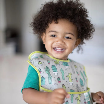 A curly-haired toddler smiles while wearing a Bumkins SuperBib®: Cacti.