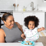 A smiling adult and child in a bright kitchen; the child points, wearing a Bumkins SuperBib®: Floral with a blue plate on a table.