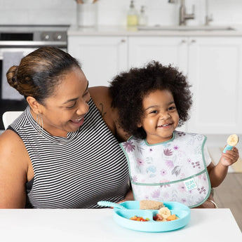 A woman smiles with a child in a Bumkins SuperBib®: Floral, holding a snack by a blue plate on a white table.