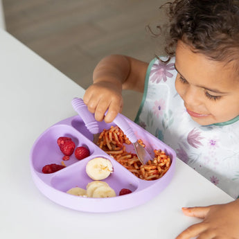A young child in a colorful Bumkins SuperBib®: Floral enjoys raspberries, spaghetti, and bananas from a purple divided plate at a table.
