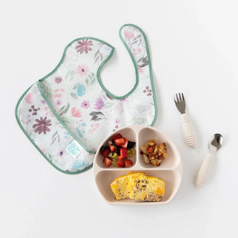 Bumkins SuperBib®: Floral lays next to utensils and sectioned plate with fruit and a cheesy omelet on a white background.