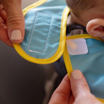 Close-up of hands fastening the hook-and-loop closure of a Bumkins SuperBib® 2 Pack: Super Mario™ around a child's neck.