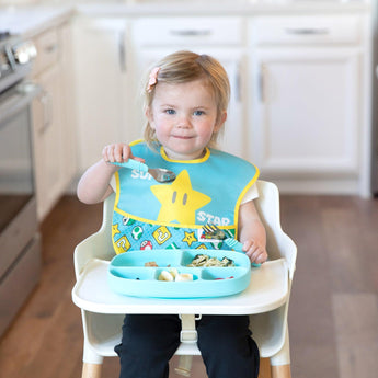 A young girl in a high chair wears a blue Bumkins SuperBib® 2 Pack: Super Mario™, holding a spoon while eating from a blue plate.