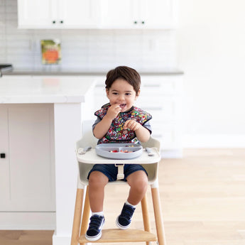 A toddler in a Bumkins SuperBib® 3 Pack: Super Mario™ Power-Up sits in a high chair, eating from a gray divided plate in a bright kitchen.