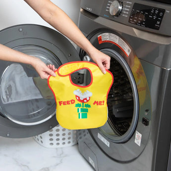 A person holds a Bumkins SuperBib® 2 Pack: Super Mario™ in front of an open front-loading washing machine.