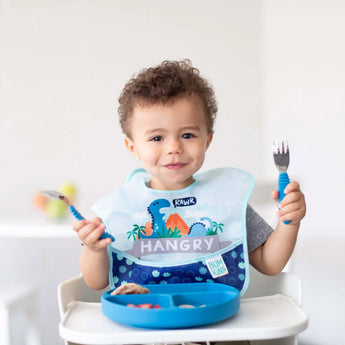 A toddler in a high chair wearing a Bumkins SuperBib® 3 Pack: Hangry, Dinosaurs, Blue Tropic eats from a blue plate with blue utensils.