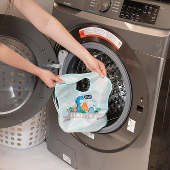A person holds a Bumkins SuperBib® 3 Pack: Hangry, Dinosaurs, Blue Tropic in front of an open front-loading washing machine.