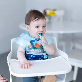 A baby in a Bumkins SuperBib® 3 Pack: Hangry, Dinosaurs, Blue Tropic sits in a high chair and chews a blue spoon.