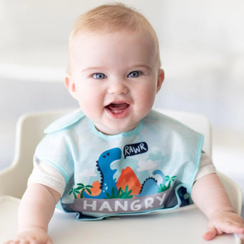 A smiling baby in a high chair wears a Bumkins SuperBib® and Sleeved Bib 3-Piece Set: Hangry, Dinosaurs, Blue Tropics.