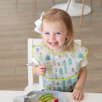 A toddler girl beams while eating with a fork, wearing a Bumkins SuperBib®: Cacti and a white bowl.