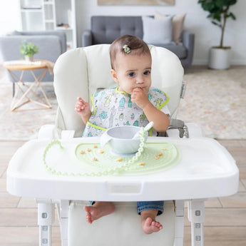 A baby sits in a high chair, wearing a Bumkins SuperBib®: Cacti and eating from a gray bowl on a light green placemat.