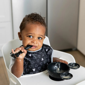 A toddler in a high chair wears a Bumkins SuperBib® 3 Pack: Love, Mickey, eating with a spoon from a black Mickey-shaped bowl.