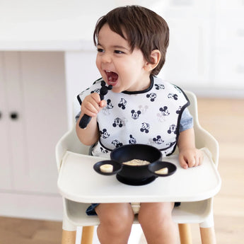 A toddler in a high chair, smiling in a Bumkins SuperBib® 3 Pack: Love, Mickey, holds a spoon while eating from a black bowl.