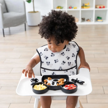 A toddler with curly hair sits in a high chair, wearing a Bumkins SuperBib® 3 Pack: Love, Mickey and eating from a Mickey Mouse-shaped plate.