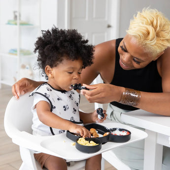An adult feeds a curly-haired toddler in a high chair wearing a Bumkins SuperBib® 3 Pack: Love, Mickey from a black Mickey-shaped plate.