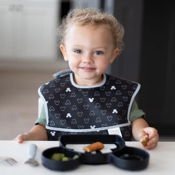 A child wearing a Bumkins SuperBib® 3 Pack: Love, Mickey smiles, ready to eat from a divided Mickey Mouse-shaped plate at a white table.