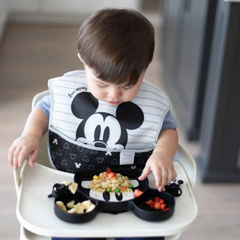 A toddler in a Bumkins SuperBib® 3 Pack: Love, Mickey enjoys a meal in a high chair with food on a black Mickey Mouse plate.