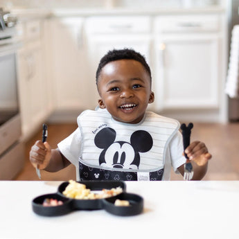 A happy child in a Bumkins SuperBib® 3 Pack: Love, Mickey sits at a white table with a Mickey-shaped plate, holding a fork and spoon.