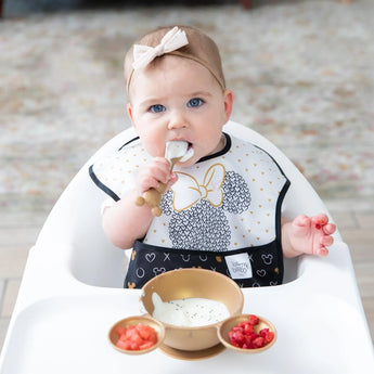 A baby in a high chair, wearing a Bumkins SuperBib® 3 Pack: Love, Minnie, eats yogurt and berries from a gold Minnie Mouse-shaped bowl.