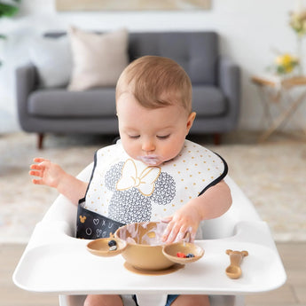 A baby is seated in a high chair, wearing a Bumkins SuperBib® 3 Pack: Love, Minnie and exploring food with their hands from a gold bowl.