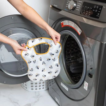 A person holds a Bumkins SuperBib® 3 Pack: Love, Minnie in front of an open front-loading washing machine.