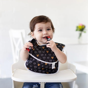 A smiling toddler in a high chair wears a Bumkins SuperBib® 3 Pack: Love, Minnie, chewing on Minnie Mouse-shaped utensils in a bright room.