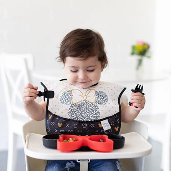 A young child sits in a high chair, wearing a Bumkins SuperBib® 3 Pack: Love, Minnie and eating from a divided plate with matching utensils.