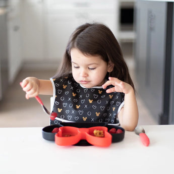 A girl wearing a Bumkins SuperBib® 3 Pack: Love, Minnie uses a fork to eat from a black & red divided Minnie Mouse plate at a white table.