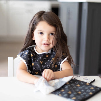 A young child with long hair sits at a white table, wearing a Bumkins SuperBib® 3 Pack: Love, Minnie and reaching into a matching pouch.