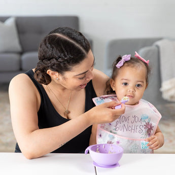 A woman in a black tank top feeds a toddler wearing a Bumkins SuperBib® 3 Pack: Love You Bunches from a purple bowl at a white table.