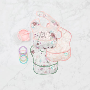 Bumkins SuperBib® 3 Pack: Love You Bunches lays next to a pink bowl and spoon set, and pastel teething rings on a marble background.