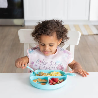A curly-haired child eats from a blue divided plate at a white table while wearing a Bumkins SuperBib® 3 Pack: Love You Bunches.