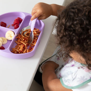 A child eats from a purple plate at a white table, wearing a Bumkins SuperBib® 3 Pack: Love You Bunches with their hand in the pocket.