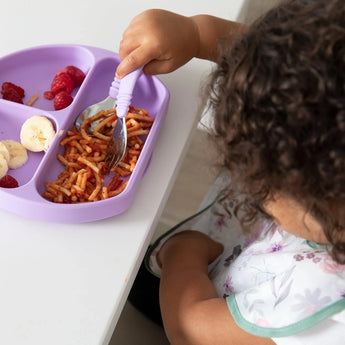 A child enjoys spaghetti, bananas, and raspberries from a purple plate on a white table, wearing a Bumkins SuperBib®: Floral.