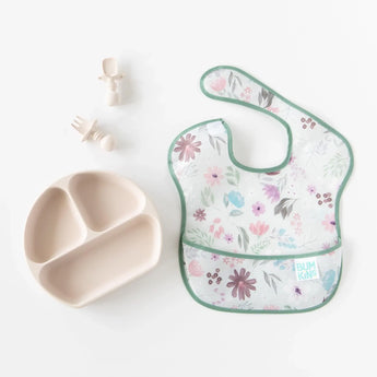 A floral Bumkins SuperBib® 3 Pack: Love You Bunches lays next to a tan divided plate and baby utensils on a white background.
