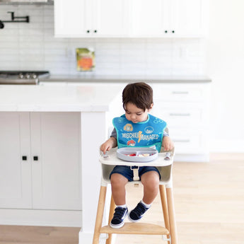 A toddler in a high chair wears a blue bib, eating fruit from a Bumkins Silicone Grip Dish: Gray in a bright, modern kitchen.