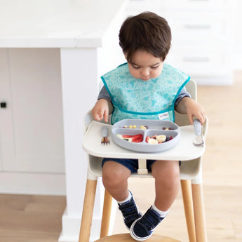 A toddler wearing a blue bib in a high chair enjoys fruit and cereal from a Bumkins Silicone Grip Dish: Gray with gray utensils.