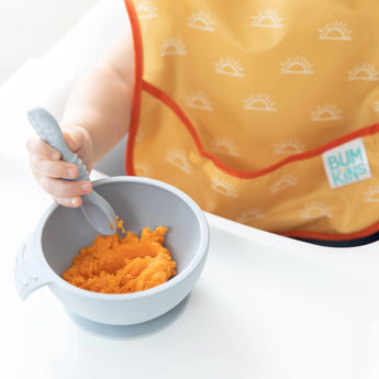 Close-up of a baby in a yellow Bumkins SuperBib® 3 Pack: Desert Boho holding a spoon over orange mash in a bowl on a white highchair tray.