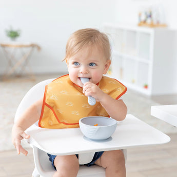 A blonde baby sits in a high chair wearing a yellow Bumkins SuperBib® 3 Pack: Desert Boho, chewing a spoon next to a bowl in a bright room.