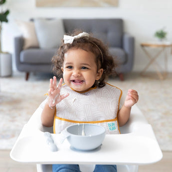 A smiling toddler in a high chair, wearing a beige Bumkins SuperBib® 3 Pack: Desert Boho, plays with food from a gray bowl.