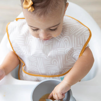 A toddler in a high chair uses a spoon to eat from a bowl, wearing a beige Bumkins SuperBib® 3 Pack: Desert Boho.
