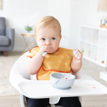 In a bright room, a baby in a high chair wearing a yellow Bumkins SuperBib® 3 Pack: Desert Boho eats from a bowl while holding a spoon.