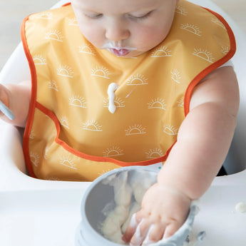 A baby in high chair wears a yellow Bumkins SuperBib® 3 Pack: Desert Boho, dripping yogurt down the front as they eat from a bowl.