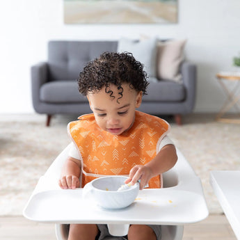 A toddler in an orange Bumkins SuperBib® 3 Pack: Desert Boho eats happily in a high chair, with a cozy living room in the background.