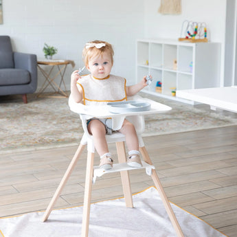 A baby in a high chair, wearing a beige Bumkins SuperBib® 3 Pack: Desert Boho, eats from a gray plate with utensils in a bright living room.