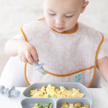 A toddler enjoys scrambled eggs with a gray fork from a gray plate, wearing a beige Bumkins SuperBib® 3 Pack: Desert Boho.