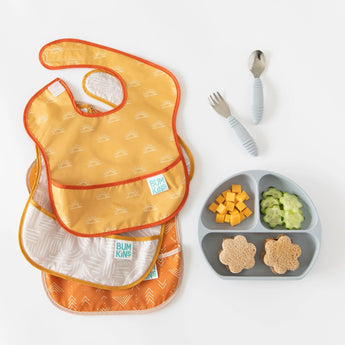 Bumkins SuperBib® 3 Pack: Desert Boho lays overlapped on a white background next to a gray divided plate of food and gray utensils.