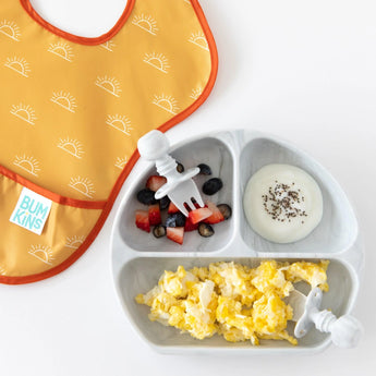 A yellow Bumkins SuperBib® 3 Pack: Desert Boho lays beside a plate of scrambled eggs, fruit, and yogurt, and utensils on a white background.