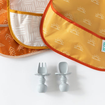 Close-up of Bumkins SuperBib® 3 Pack: Desert Boho arranged on a white background with gray baby utensils underneath.