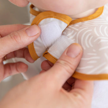 Close-up of hands fastening the hook-and loop closure of a beige Bumkins SuperBib® 3 Pack: Desert Boho around a baby's neck.